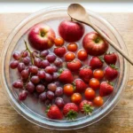 Mixed fruit and vegetables including apples, grapes, strawberries, and cherry tomatoes soaking in a baking soda fruit wash solution in a large glass bowl