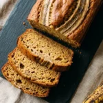 Sliced homemade banana bread on a dark slate board showing moist golden crumb, with ripe bananas alongside