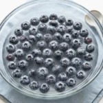 Fresh blueberries soaking in a baking soda water solution in a shallow clear bowl, showing the silvery bloom on the berry skins
