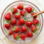 Fresh whole strawberries with tops on soaking in a baking soda water solution in a clear glass bowl