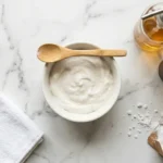 Overhead flat-lay of baking soda paste in a white ceramic bowl surrounded by cleaning tools on a marble countertop