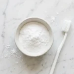 A small glass bowl of white baking soda powder next to a soft-bristled toothbrush on a clean white marble surface