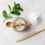 Glass jar of homemade baking soda toothpaste paste with a wooden spoon, fresh peppermint, and a soft toothbrush on white marble