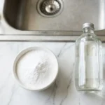 Baking soda and white vinegar next to a kitchen sink drain ready for cleaning
